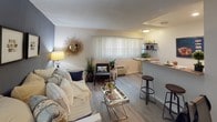 This single-floor unit is thoughtfully designed to maximize functionality within a compact footprint. The open-concept layout connects the living area, kitchen, and entryway, creating a cohesive and efficient flow throughout the space. Wood-look plank flooring runs continuously across the main areas, lending a warm, unified aesthetic.

The living area serves as the central hub, anchored by a bold navy-blue accent wall that adds depth and contrast against the neutral white walls. A large window with vertical blinds allows natural light to fill the space, while a dedicated desk nook provides a practical workspace. The area is versatile enough to accommodate both a seating arrangement and a workspace simultaneously.

The kitchen runs along one side of the open layout and features white cabinetry, speckled countertops, a gas range, refrigerator, and a raised breakfast bar with seating. Open shelving above the bar offers additional display and storage options.

The bathroom is clean and well-appointed, featuring an extended vanity with a large mirror, white cabinetry, speckled countertops, and a separate water closet for added privacy. Light-toned flooring and bright overhead lighting contribute to an airy, functional feel throughout.