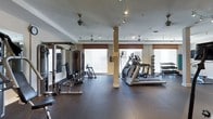 This fitness center offers a comprehensive workout environment designed to accommodate a variety of exercise routines. The space is outfitted with a solid selection of strength training equipment, including cable machines and weight racks, alongside cardio machines such as ellipticals and treadmills, all arranged to allow comfortable movement throughout the floor. Durable rubber flooring covers the entire workout area, providing both safety and cushioning underfoot. Structural columns are integrated into the layout without impeding the functional use of the space. Large windows dressed with blinds and decorative valances line the walls, inviting an abundance of natural light and lending the room a bright, welcoming atmosphere. Multiple ceiling fans are positioned throughout to ensure consistent airflow during workouts, complemented by a track lighting system that delivers focused, adjustable illumination. The overall design balances practicality with a polished aesthetic, making it a well-appointed facility suited for a full range of fitness activities.