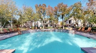 This community amenity space centers around a generously sized outdoor pool area, designed to evoke a resort-like atmosphere. The pool features vivid turquoise water and a raised spa with tiled surrounds, seamlessly integrated into the design. Surrounding the pool deck are multiple lounge chairs and shaded umbrella seating areas, ideal for relaxing outdoors. Mature trees with seasonal foliage frame the space, providing natural shade and a scenic backdrop, while multi-story residential buildings border the perimeter, creating an enclosed, private setting. A grill station is also visible within the outdoor area, adding to its recreational appeal.

The fitness center is a well-organized, functional space equipped with a range of cardio machines, including treadmills and ellipticals from recognized brands such as Precor and Matrix. Strength training equipment, including a cable machine and free weights, is also available. Full-length mirrors line one wall, enhancing the sense of space and allowing users to monitor their form. Multiple wall-mounted televisions and a wall clock add to the practicality of the room. Natural light enters through horizontal-slatted blinds on multiple windows, complemented by overhead fluorescent lighting, creating a bright and energetic workout environment.