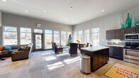 This clubhouse in Colorado Springs, Colorado, offers a thoughtfully designed single-floor layout that combines a fully equipped kitchen with a generous living area. The open-concept floor plan creates a welcoming, communal atmosphere ideal for gatherings and everyday use.

The living area is bathed in natural light, courtesy of large floor-to-ceiling windows that line the exterior walls and provide views of the surrounding outdoor space. The soaring ceilings amplify the sense of openness, while neutral gray walls and patterned carpet flooring lend a modern, polished aesthetic. Comfortable seating arrangements, a wall-mounted flat-screen TV, and glass-top coffee tables make this space both functional and inviting.

The kitchen is well-equipped with dark cabinetry, stainless steel appliances including a refrigerator, dishwasher, range, and microwave, along with a subway tile backsplash and a central island with counter seating. A dedicated beverage station is conveniently positioned nearby, adding to the space's hosting capabilities.

Recessed lighting throughout complements the abundant daylight, ensuring the space remains bright and functional at all times. Direct access to an outdoor area is provided through a glass-panel entry door, seamlessly connecting the interior with the exterior environment.