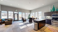 This clubhouse in Colorado Springs, Colorado, offers a thoughtfully designed single-floor layout that combines a fully equipped kitchen with a generous living area. The open-concept floor plan creates a welcoming, communal atmosphere ideal for gatherings and everyday use.

The living area is bathed in natural light, courtesy of large floor-to-ceiling windows that line the exterior walls and provide views of the surrounding outdoor space. The soaring ceilings amplify the sense of openness, while neutral gray walls and patterned carpet flooring lend a modern, polished aesthetic. Comfortable seating arrangements, a wall-mounted flat-screen TV, and glass-top coffee tables make this space both functional and inviting.

The kitchen is well-equipped with dark cabinetry, stainless steel appliances including a refrigerator, dishwasher, range, and microwave, along with a subway tile backsplash and a central island with counter seating. A dedicated beverage station is conveniently positioned nearby, adding to the space's hosting capabilities.

Recessed lighting throughout complements the abundant daylight, ensuring the space remains bright and functional at all times. Direct access to an outdoor area is provided through a glass-panel entry door, seamlessly connecting the interior with the exterior environment.