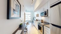 This single-floor unit showcases a contemporary design with a clean, neutral palette throughout. Light hardwood flooring extends across the entire space, contributing to a cohesive and airy feel. The galley-style kitchen is a standout feature, lined with flat-front white cabinetry, subway tile backsplash, and dark quartz countertops. Stainless steel appliances — including a range, over-the-range microwave, and dishwasher — are complemented by under-cabinet lighting and a stainless undermount sink. Track lighting along the kitchen ceiling adds a modern, functional touch.

Adjacent to the kitchen, a built-in desk area serves as a compact workspace or dining nook, with bar-height seating. The living area features notably tall ceilings, a ceiling fan, and a large window that draws in natural light. A sliding door closet with built-in shelving provides practical storage. The bedroom area flows openly from the main living space, maintaining the unit's open-concept character.

The bathroom is finished with a white vanity, dark countertop, undermount sink, a large mirror with modern vanity lighting, and ample drawer storage — consistent with the unit's modern, minimalist aesthetic throughout.