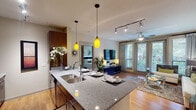 This single-story 2-bedroom, 2-bathroom residence in Houston, TX offers a thoughtfully designed open-concept layout that integrates the kitchen, dining, and living areas into one cohesive space. The kitchen is outfitted with rich dark-wood cabinetry extending to the ceiling, stainless steel appliances including a refrigerator, electric range, and microwave, and a large granite-topped island with an undermount double sink. Vibrant yellow pendant lights illuminate the island, which provides seating for multiple bar stools. Track lighting runs throughout the kitchen and into the living area, complementing the ceiling fan overhead. The living room features hardwood-style flooring, a wall-mounted flat-screen TV, and floor-to-ceiling glass doors that open to a private outdoor space with lush greenery views. Both bedrooms are carpeted and receive generous natural light through large windows with views of surrounding greenery, each equipped with a ceiling fan. The two bathrooms are finished in neutral tones, each featuring a tub-shower combination with dark cabinetry for storage. A stacked in-unit washer and dryer is conveniently located within the unit, adding everyday practicality to this well-finished Houston residence.