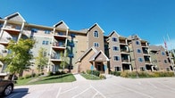 This fitness center is housed within a multi-story residential building, identifiable by its distinctive combination of tan brick, green and beige siding, and multiple private balconies framed against a clear sky. The building's welcoming entrance is marked by a gabled canopy and well-maintained landscaping.

Inside, the fitness center offers a practical and functional layout designed to accommodate a range of workout routines. The space features a dark rubberized floor that provides durability and comfort underfoot. Cardio equipment includes treadmills and an elliptical trainer from reputable brands, positioned to face a wall-mounted flat-screen television and natural light from several windows fitted with horizontal blinds. Strength training is supported by a multi-station cable machine, while exercise balls add versatility for stretching and core work.

A large floor-to-ceiling mirror spans one wall, visually expanding the room and allowing users to monitor their form during exercise. The room is lit by recessed fluorescent ceiling fixtures, ensuring consistent, bright illumination throughout. Convenience amenities include a stainless steel drinking fountain, a wall-mounted paper towel and sanitizing spray dispenser, and a thermostat for climate control. A secured entry door with an automatic closer ensures controlled access to the space.