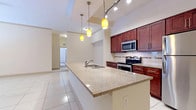 This single-floor residence offers a thoughtfully designed open layout that seamlessly connects the kitchen and living areas. The kitchen is a standout feature, centered around a generous granite-topped island with an undermount sink, complemented by rich dark wood cabinetry, a decorative tile backsplash, and a full suite of stainless steel appliances including a refrigerator, electric range, and built-in microwave. Warm pendant lighting above the island adds a stylish touch, while track lighting illuminates the space further. Large-format white tile flooring extends throughout the main living and kitchen areas, lending a clean, bright aesthetic.

The bedrooms are carpeted for comfort and receive natural light through window blinds. The primary bedroom provides direct access to a well-appointed en-suite bathroom featuring a dual-sink vanity with granite countertops, rich wood cabinetry, a large mirror, and tile flooring. A spacious walk-in closet with wire shelving offers generous storage capacity.

A dedicated laundry area houses a top-loading washer with shelf storage above. Throughout the residence, the neutral color palette, consistent finishes, and practical layout combine to create a cohesive and functional living environment.