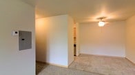 This single-floor space is designed with a straightforward, functional layout that makes efficient use of its footprint. The interior is defined by clean white walls and standard-height ceilings, contributing to a bright and neutral ambiance throughout.

A notable feature of the layout is a partial dividing wall that creates a natural separation between the carpeted living area and an adjacent zone with light tile flooring. This architectural element adds subtle definition to the space without fully closing off one area from another, maintaining an open and connected feel.

The living area is outfitted with carpet underfoot and is equipped with a ceiling fan fitted with a light fixture, providing both air circulation and overhead illumination. The transition to tile flooring marks a shift toward the kitchen area, which is partially visible beyond the dividing wall. Cabinetry and appliances can be glimpsed from this vantage point, suggesting a compact but functional kitchen space.

The overall aesthetic is clean and understated, with neutral tones across the walls, flooring, and ceiling. The space is well-suited for practical, comfortable everyday living, offering a cohesive and easy-to-maintain interior environment.