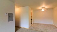 This single-floor space is designed with a straightforward, functional layout that makes efficient use of its footprint. The interior is defined by clean white walls and standard-height ceilings, contributing to a bright and neutral ambiance throughout.

A notable feature of the layout is a partial dividing wall that creates a natural separation between the carpeted living area and an adjacent zone with light tile flooring. This architectural element adds subtle definition to the space without fully closing off one area from another, maintaining an open and connected feel.

The living area is outfitted with carpet underfoot and is equipped with a ceiling fan fitted with a light fixture, providing both air circulation and overhead illumination. The transition to tile flooring marks a shift toward the kitchen area, which is partially visible beyond the dividing wall. Cabinetry and appliances can be glimpsed from this vantage point, suggesting a compact but functional kitchen space.

The overall aesthetic is clean and understated, with neutral tones across the walls, flooring, and ceiling. The space is well-suited for practical, comfortable everyday living, offering a cohesive and easy-to-maintain interior environment.