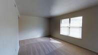 This single-floor residence offers a practical and straightforward layout designed for comfortable everyday living. The galley-style kitchen is well-equipped, featuring warm wood cabinetry with ample upper and lower storage, a white gas range, a full-size refrigerator, and a double-basin sink with a window view. A fluorescent overhead light provides strong task lighting, while a ceiling fan positioned at the kitchen's entrance ensures good airflow throughout the space. The tile flooring in the kitchen is durable and easy to maintain, and the layout allows for efficient movement between appliances and prep areas. Beyond the kitchen, the living area transitions to plush carpeted flooring, creating a softer, more comfortable atmosphere. A large window fitted with horizontal blinds allows natural light to fill the room, casting a warm glow across the neutral-toned walls and light carpet. The space is clean and open, offering flexibility for a variety of furniture arrangements. The overall design is simple and functional, with a neutral color palette that provides a versatile backdrop for personalization. Consistent finishes and a well-considered floor plan make this a cohesive and livable space.
