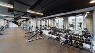 Located at 770 Jackson Ave in Hoboken, New Jersey, this contemporary two-floor fitness center offers a comprehensive and well-equipped environment designed to support a wide range of workout routines. The facility features an industrial-modern aesthetic, with striking black exposed ceilings, linear LED lighting, polished concrete columns, and durable rubber flooring throughout.

The cardio zone is generously outfitted with a full row of Life Fitness treadmills positioned along floor-to-ceiling windows, providing views of the surrounding Hoboken cityscape. Additional cardio options include Peloton spin bikes, elliptical machines, stair climbers, and rowing machines, accommodating both high-intensity and low-impact training preferences.

The strength training area is equally well-appointed, featuring multiple weight benches, cable machines, squat racks, and an extensive dumbbell rack lining a full-length mirrored wall that enhances the sense of space. A dedicated functional training zone offers medicine balls, stability equipment, and open floor space for dynamic exercises.

A separate enclosed room provides a quieter setting for elliptical and rowing machine use. The facility also includes locker storage, a water fountain station, and staircase access between floors. A decorative mural adds an artistic focal point near the treadmill area, contributing to the space's polished and energetic atmosphere.