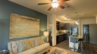 This single-story space showcases an open-concept design that seamlessly connects the living area and kitchen into one cohesive, functional environment. The living area is anchored by a bold teal accent wall adorned with a large-scale abstract painting, adding a striking focal point to the room. A ceiling fan with a light fixture overhead provides both airflow and ambient illumination, complementing the warm tones throughout the space. The seating area features a tufted cream sofa accented with decorative patterned pillows, contributing to a welcoming and contemporary atmosphere.

A structural column elegantly separates the living and kitchen zones while maintaining the open flow between them. The kitchen is outfitted with dark cabinetry, open shelving, a light-toned countertop, and a breakfast bar with seating, making it ideal for casual dining and entertaining. Track lighting above the kitchen ensures the workspace is well-lit. Dark hardwood-style flooring runs throughout, grounding the design and tying both areas together. The overall aesthetic balances modern sophistication with comfortable, livable style.