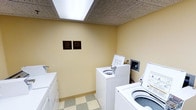 This shared laundry facility is a compact, single-floor space designed for functional, everyday use. The room features light yellow walls that create a clean, neutral environment, complemented by a classic drop ceiling with recessed fluorescent lighting panels that ensure the area is well-lit. The flooring consists of a checkered vinyl tile in muted tones, adding a practical and easy-to-maintain surface underfoot. Several top-loading washing machines and dryers are arranged along the walls, providing multiple units for simultaneous use. A payment station is mounted on the wall, indicating coin-operated or card-operated functionality. Two informational plaques are displayed prominently on the wall, outlining usage rules and management contact information for residents. A utility shelf or counter is partially visible along one side, offering a convenient surface for sorting or folding laundry. The overall ambiance is straightforward and utilitarian, prioritizing practicality and ease of use for building occupants.