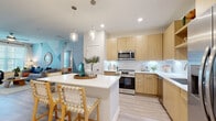 This single-floor residence offers a thoughtfully designed layout across two bedrooms and two bathrooms, providing a comfortable and functional living environment. The open-concept kitchen and living area serves as the heart of the home, where light wood flat-front cabinetry, a white quartz island with bar seating, pendant lighting, and stainless steel appliances—including a French door refrigerator, range, and microwave—create a modern, cohesive aesthetic. Light wood-tone flooring runs throughout, unifying the space.

The living room features a ceiling fan, large windows with abundant natural light, and a screened balcony with views of landscaped grounds and palm trees—ideal for relaxed outdoor seating. A geometric accent wall in soft blue tones adds visual interest to the living area.

Both bedrooms are comfortably proportioned, each equipped with ceiling fans, large windows, and light-filled interiors. The primary bedroom connects to a spacious walk-in closet outfitted with multi-tiered wire shelving and hanging rods.

The bathrooms feature light wood vanities with white quartz countertops and marble-style tiled tub/shower surrounds. Additional conveniences include a dedicated laundry area and hallway connecting all rooms efficiently.