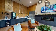 This single-floor community lounge is designed to offer a welcoming and functional gathering environment. The space is anchored by a grand open area with soaring double-height ceilings, wide-plank hardwood flooring, and an abundance of natural light streaming through oversized windows. Distinct seating zones are arranged throughout, including a conversation area with plush gray sofas and a dining table with leather chairs, creating versatile spaces for socializing or quiet relaxation.

A dedicated lounge nook features a sleek dark fireplace clad in tile, flanked by a wall-mounted television and upholstered built-in banquette seating. Cognac leather armchairs and teal accent pillows add warm, contemporary color throughout. Adjacent to this area, a well-appointed coffee bar runs along a wood-paneled accent wall, complete with dark navy cabinetry, a white countertop, pendant lighting, and café-style decorative elements.

High windows in the nook provide views of the surrounding landscape, enhancing the connection to the outdoors. Textural contrasts are a defining design element, with herringbone tile accents, wood wall paneling, concrete-look surfaces, and curated wall art contributing to the space's modern yet inviting character. The overall ambiance is refined and comfortable, well-suited for both casual gatherings and everyday use.