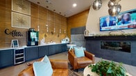 This single-floor community lounge is designed to offer a welcoming and functional gathering environment. The space is anchored by a grand open area with soaring double-height ceilings, wide-plank hardwood flooring, and an abundance of natural light streaming through oversized windows. Distinct seating zones are arranged throughout, including a conversation area with plush gray sofas and a dining table with leather chairs, creating versatile spaces for socializing or quiet relaxation.

A dedicated lounge nook features a sleek dark fireplace clad in tile, flanked by a wall-mounted television and upholstered built-in banquette seating. Cognac leather armchairs and teal accent pillows add warm, contemporary color throughout. Adjacent to this area, a well-appointed coffee bar runs along a wood-paneled accent wall, complete with dark navy cabinetry, a white countertop, pendant lighting, and café-style decorative elements.

High windows in the nook provide views of the surrounding landscape, enhancing the connection to the outdoors. Textural contrasts are a defining design element, with herringbone tile accents, wood wall paneling, concrete-look surfaces, and curated wall art contributing to the space's modern yet inviting character. The overall ambiance is refined and comfortable, well-suited for both casual gatherings and everyday use.