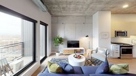 This single-floor unit showcases a compelling blend of industrial and contemporary design, anchored by exposed concrete ceilings that extend throughout every room. The living area is a standout space, featuring large black-framed windows and a sliding glass door that open to a private balcony with sweeping city views. Light hardwood flooring runs throughout the main living spaces, complementing the raw concrete overhead and creating a warm contrast to the industrial aesthetic. Exposed ductwork adds to the urban character of the room.

The kitchen is well-appointed with flat-front cabinetry in a light wood tone, stainless steel appliances including a full-size refrigerator, gas range, microwave, and dishwasher, paired with white countertops and a subway tile backsplash. A kitchen island provides additional workspace and casual seating.

The bedroom continues the concrete ceiling detail, offering city views through black-framed windows and a sliding barn door for privacy. A dedicated picture ledge adds a curated display element above the bed.

The bathroom features a double-sink vanity with a light wood base, a large LED-backlit mirror, white countertops, and a glass-enclosed walk-in shower, all finished in a clean, modern palette.