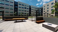 Located at 87 Union St in New Haven, Connecticut, this outdoor courtyard is a well-designed communal space nestled within a contemporary multi-story residential complex. The building's exterior showcases a modern architectural style, combining gray horizontal siding, large windows, and black metal accents that frame the courtyard on multiple sides, creating an intimate yet open-air atmosphere.

The patio level is finished with large-format light stone tiles that lend a clean, polished look underfoot. The space is thoughtfully furnished with warm wood-and-metal benches positioned throughout, offering comfortable areas to gather and relax. A central rectangular fire pit serves as a focal point, making the courtyard an inviting setting during cooler evenings. Raised planters with low stone-clad walls topped with granite-style caps define the edges of the space and introduce greenery that softens the hardscape.

String lights are strung between tall black poles, adding an ambient glow for evening use. Steps with metal railings lead to an adjacent landscaped lawn area, expanding the usable outdoor space. Glass-panel entry doors with dark metal frames provide access to interior amenities, and the surrounding balconies overlook the courtyard, reinforcing the connected, community-oriented design. The overall ambiance is modern, clean, and welcoming.