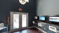 Located in Dallas, this single-floor communal space combines modern aesthetics with practical amenities. The interior is defined by striking soaring ceilings that amplify the sense of openness, finished in a sophisticated deep slate-blue that lends the space a contemporary, polished character. Rich dark hardwood flooring grounds the room and contrasts elegantly with the crisp white architectural details throughout.

A dedicated business center area features a sleek countertop workstation equipped with multiple desktop computers and white modern bar-height seating, offering a functional space for productivity. Overhead, a bold floral-inspired chandelier serves as a statement fixture, adding an artistic touch to the otherwise clean-lined interior.

Large glass double doors flood the space with natural light and provide direct views and access to an outdoor area, creating a seamless connection between the interior and exterior environments. The overall ambiance is professional yet inviting, blending high-design elements with everyday utility. This thoughtfully designed space reflects a commitment to both form and function, making it well-suited as a shared amenity or community hub.