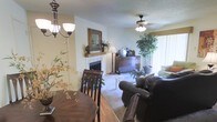 This single-floor space offers a welcoming and functional layout that seamlessly connects the dining and living areas. The living room is anchored by a classic white-brick fireplace adorned with a decorative mirror and mantel accents, creating a cozy focal point. A ceiling fan with a light fixture provides comfort overhead, while plush carpeting underfoot adds warmth to the space. The living area is furnished with a large leather sofa and complemented by lush indoor plants that bring a touch of natural greenery indoors. A wooden credenza positioned near the window serves as both a storage and display surface. Large windows fitted with vertical blinds allow generous natural light to fill the room, enhancing the bright and airy ambiance. Transitioning into the dining area, hardwood-style flooring anchors a dark wood dining table and chairs beneath a charming chandelier, creating an intimate space for meals. The overall aesthetic balances comfort and classic design, with warm tones, soft textures, and carefully chosen furnishings that contribute to a cohesive and livable environment throughout.