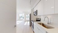 This thoughtfully designed single-floor unit showcases a clean, contemporary aesthetic defined by an all-white palette, light hardwood flooring, and an abundance of natural light. The open-concept layout maximizes every square foot, creating a seamless flow between the kitchen and living area.

The kitchen is a standout feature, outfitted with high-gloss white cabinetry extending to the ceiling, a white tile backsplash, light quartz countertops, and a full suite of stainless steel appliances including a refrigerator, electric range, built-in microwave, and dishwasher. A deep stainless steel sink with a modern pull-down faucet completes the functional workspace. Track lighting overhead ensures ample task illumination.

The open living area is bright and airy, benefiting from large floor-to-ceiling windows that frame outdoor views and flood the space with natural sunlight. Light wood-toned flooring runs continuously throughout, enhancing the sense of openness.

The bathroom maintains the same minimalist design language, featuring floor-to-ceiling white tile surround, a soaker tub with an overhead shower, chrome fixtures, a floating white vanity, and coordinating wood-tone flooring. Every detail reflects a modern, move-in-ready finish with a cohesive and understated style throughout.