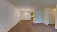 This single-floor space offers a straightforward and functional layout designed to maximize usability. The main living and dining area is open and spacious, featuring neutral white walls that create a clean, bright backdrop. Wall-to-wall carpeting in a warm beige tone covers the living area, contributing a sense of comfort underfoot. A modest chandelier hangs centrally, providing a subtle decorative focal point while illuminating the dining space below.

The kitchen is accessible directly from the main living area, separated by a partial wall that defines the space without fully enclosing it. Inside the kitchen, warm medium-toned wood cabinetry lines the upper and lower walls, offering ample storage. The kitchen floor transitions from the carpeted living area to a hard surface, providing a practical and easy-to-clean workspace.

The overall color palette throughout the space is understated and neutral, with white walls and ceilings creating a sense of openness. The layout is efficient and cohesive, making the space well-suited for everyday living. The single-floor configuration ensures ease of movement throughout, with all areas conveniently accessible from one level.