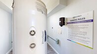 This single-room tanning facility is a compact, efficiently arranged hallway space designed specifically for UV tanning services. The room is finished with soft light gray walls and durable wood-look luxury vinyl plank flooring, creating a clean and calming atmosphere. A ProSun tanning bed is the centerpiece of the space, positioned prominently within the room and accompanied by all necessary operational equipment. A wall-mounted paper towel dispenser provides convenient access to sanitation supplies, supporting hygiene standards within the facility. Detailed tanning rules and UV radiation safety signage from The District are clearly posted on the wall, ensuring clients are fully informed prior to use. A support handrail is affixed to one wall, adding a practical safety feature. Rustic wooden crate shelving built into one wall offers storage for tanning products, while a gold-framed decorative piece adds a touch of style to the otherwise utilitarian space. Multiple electrical outlets and control panels are distributed along the walls to accommodate the equipment's power needs. A window allows a measure of natural light into the space, contributing to a bright and welcoming feel. Overall, the room is a purpose-built, well-organized tanning suite that balances function with clean, modern aesthetics.