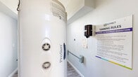 This single-room tanning facility is a compact, efficiently arranged hallway space designed specifically for UV tanning services. The room is finished with soft light gray walls and durable wood-look luxury vinyl plank flooring, creating a clean and calming atmosphere. A ProSun tanning bed is the centerpiece of the space, positioned prominently within the room and accompanied by all necessary operational equipment. A wall-mounted paper towel dispenser provides convenient access to sanitation supplies, supporting hygiene standards within the facility. Detailed tanning rules and UV radiation safety signage from The District are clearly posted on the wall, ensuring clients are fully informed prior to use. A support handrail is affixed to one wall, adding a practical safety feature. Rustic wooden crate shelving built into one wall offers storage for tanning products, while a gold-framed decorative piece adds a touch of style to the otherwise utilitarian space. Multiple electrical outlets and control panels are distributed along the walls to accommodate the equipment's power needs. A window allows a measure of natural light into the space, contributing to a bright and welcoming feel. Overall, the room is a purpose-built, well-organized tanning suite that balances function with clean, modern aesthetics.