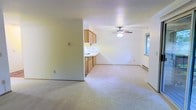 This single-floor space offers a practical and straightforward layout designed for comfortable everyday living. The open floor plan seamlessly connects the living area to the kitchen, creating a sense of spaciousness throughout. The living area is finished with neutral wall-to-wall carpeting, which adds a warm and quiet underfoot feel, while the light-colored walls contribute to a bright and airy ambiance. A ceiling fan with an integrated light fixture is centrally positioned overhead, providing both air circulation and overhead illumination. Natural light filters in through a window on the side wall, offering a view of surrounding greenery outside. The kitchen area is efficiently arranged with warm-toned wood cabinetry above and below the counter, providing ample storage and workspace. A built-in sink and countertop complete the functional kitchen setup. A baseboard heater runs along the far wall, ensuring climate control throughout the space. The overall design is clean and understated, with simple finishes that lend themselves well to personalization. This space provides a comfortable, functional foundation with an efficient single-level layout suited to a variety of living needs.