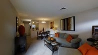 This single-story residence features an open-concept floor plan that seamlessly connects the main living areas, creating a cohesive and functional layout. The living room serves as the central gathering space, furnished with a plush gray sofa accented by rust-colored throw pillows and anchored by a dark metal and glass coffee table. A large framed artwork with warm earth tones adds visual interest to the neutral walls. Soft carpeting extends throughout the living area, contributing to a comfortable and inviting atmosphere.

The dining area flows naturally from the living room and is illuminated by a classic chandelier, providing warm ambient lighting over the round dining table and set of dark wood chairs. Decorative accents, including a table centerpiece and a floor lamp, enhance the warmth of the space. Beyond the dining area, the kitchen is visible through an open pass-through or partial wall, featuring light cabinetry and a bright, functional workspace. A second ceiling fan light fixture in this zone ensures the connected areas remain well-lit. The overall design presents a practical, warm, and welcoming single-level home.