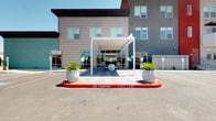 This single-story leasing office presents a polished, contemporary design that makes an immediate impression from the exterior. The building features a clean facade with horizontal lap siding in neutral gray tones, floor-to-ceiling glass windows, and a striking white pergola-style canopy over the main entrance. The paved entryway is accented with large decorative planters, creating an inviting arrival experience.

Inside, the open-plan interior is thoughtfully designed for both comfort and functionality. The main lounge area centers around a curated seating arrangement of bold teal upholstered chairs surrounding a round wood-topped coffee table, all set atop a geometric multicolored area rug over light wood-look flooring. A statement chandelier featuring an asymmetric branching frame with globe bulbs serves as a focal point overhead, complementing the recessed lighting throughout.

The space flows seamlessly into additional areas, including what appears to be a refreshment station with dark cabinetry along one wall, offering a hospitality-forward touch. Structural columns define zones without interrupting the openness of the space, while large windows allow natural light to fill the interior and provide views of the surrounding landscape.