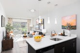 This single-floor residence showcases a thoughtfully designed open layout with light wood-style flooring throughout the main living areas. The hallway makes an immediate impression with a striking metal sliding barn door mounted on a black track, offering both functional room division and an industrial-modern design element.

The living area is bright and welcoming, bathed in natural light from large sliding glass doors that open to a private outdoor patio. The seamless indoor-outdoor connection enhances the sense of space and brings in views of the surrounding greenery.

The kitchen features dark wood cabinetry, stainless steel appliances including a range, microwave, and refrigerator, and a white countertop peninsula with bar seating beneath pendant lighting — ideal for casual dining or entertaining.

The bedroom is a comfortable, well-proportioned retreat with neutral tones, large windows fitted with blinds and curtains, a closet, and soft carpet underfoot.

The bathroom is finished with dark wood-toned cabinetry, a wide vanity with a single sink, a large mirror, and light wood-style tile flooring, creating a clean and modern aesthetic throughout.