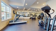 Located at 31 Walter Ave in Highland Park, NJ, this fitness center is a thoughtfully designed, single-floor facility built to accommodate a comprehensive workout experience. The space is defined by a bold red and white color scheme, with vibrant red accent walls contrasting cleanly against white surfaces and structural columns throughout.

The main workout floor is generously proportioned, finished with durable dark rubber flooring that provides traction and impact absorption across all exercise zones. Large wall-mounted mirrors span multiple sections of the room, visually expanding the space while allowing users to monitor their form during training sessions.

The strength training area is well-appointed with cable machines, multi-functional weight stations, adjustable benches, and a full rack of kettlebells and free weights. The cardio zone features multiple treadmills and an elliptical machine, all positioned beneath wall-mounted flat-screen televisions for entertainment during workouts. Stability balls and resistance accessories are also available for functional training.

Recessed overhead lighting ensures the entire floor is evenly and brightly illuminated. The facility includes separate entry and interior doors, with clearly marked emergency exits. The clean, organized layout offers ample open floor space between equipment stations, making the environment accessible and easy to navigate for users of all fitness levels.