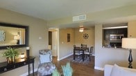This single-floor residence offers a thoughtfully designed layout that emphasizes open, connected living spaces. The living and dining areas flow seamlessly into one another, creating a cohesive and inviting atmosphere. Rich hardwood flooring runs throughout, anchoring the space with warmth and a sense of continuity.

The living area is bright and comfortable, featuring neutral beige walls that lend a calm, versatile backdrop for furnishings. A patterned accent chair adds a touch of character, while decorative indoor plants introduce a refreshing natural element. Ceiling-mounted HVAC vents indicate a centralized climate control system for year-round comfort.

The dining area is defined by a classic dark wood table and matching ladder-back chairs, set atop a geometric area rug that adds subtle visual interest underfoot. A ceiling fan with a light fixture overhead provides both air circulation and ambient lighting. Decorative wall accents lend personality without overwhelming the clean, understated aesthetic.

A hallway visible from the living area leads to additional rooms, hinting at a practical and well-organized floor plan. The white panel doors and bright white baseboards provide crisp contrast against the warm wall tones. Overall, the space presents a tidy, functional home with a welcoming ambiance suited for everyday living.
