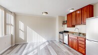 Located at 1845 Summit Pl NW in Washington, DC, this efficiently designed single-floor unit offers a bright and functional layout ideal for urban living. The open living area is flooded with natural light through large windows dressed with vertical blinds, casting dramatic light patterns across the light-toned hardwood-style flooring that extends throughout the entire space.

The kitchen is integrated into the main living area and features warm cherry wood cabinetry, a gas range, dishwasher, stainless steel refrigerator, microwave, and a sink with ample counter space — providing all essential cooking amenities in a compact, well-organized footprint. Flush-mount ceiling light fixtures illuminate both the kitchen and living zones evenly.

The bathroom is finished with neutral beige tile surrounding a full soaking tub and shower combination, complemented by a pedestal-style sink, a wall mirror, and shelving for storage. The flooring features classic white tile, maintaining a clean and functional aesthetic.

Additional features include a dedicated closet for storage, a visible electrical panel, and baseboard heating units. The crisp white walls and warm flooring create a bright, welcoming ambiance throughout. The unit's location in the lively Columbia Heights/Adams Morgan corridor of Washington, DC, provides convenient access to dining, transit, and city amenities.