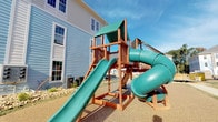 This outdoor playground is a well-equipped recreational space designed to provide an engaging environment for children within a modern residential community. The centerpiece of the playground is a sturdy, multi-level wooden play structure constructed from rich, warm-toned timber framing. The structure features a covered upper platform with a green canopy roof, offering shade and an elevated vantage point.

Two distinct slide options descend from the platform: a broad, open straight slide ideal for quick descents, and an enclosed spiral tube slide that adds an element of adventure. A tire swing is suspended beneath the main deck, adding another interactive play element. The entire structure is anchored on a rubberized, impact-absorbing surface that provides both safety and durability underfoot.

The playground is enclosed by a black metal railing fence, clearly defining the play area within the larger community grounds. Manicured green lawns, concrete walkways, and multi-story residential buildings with balconies and siding facades frame the surroundings, reflecting the well-maintained character of the neighborhood. The open-sky setting ensures ample natural light throughout the day, making this playground a bright and inviting destination for outdoor activity.