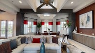This single-floor community space makes an immediate impression through its grand entrance foyer, where floor-to-ceiling black-framed windows flank a set of glass double doors, flooding the entryway with natural light. Light wood-look tile flooring and built-in bench seating establish a welcoming, contemporary tone from the moment of arrival.

The interior opens into a spacious main gathering area defined by soaring vaulted ceilings adorned with bold, dark reclaimed wood beam detailing that forms a striking architectural centerpiece overhead. Dark textured columns frame the open-plan layout, creating natural zones between the lounge and dining areas without sacrificing the sense of openness.

The lounge area is anchored by a wall-mounted linear fireplace set into a floor-to-ceiling textured tile surround, complemented by a large flat-screen display above. Accent walls in deep burgundy add warmth and visual contrast against the light grey walls and neutral flooring throughout. Bold red-and-white drapery panels dress the abundant windows, which provide views to the surrounding outdoor landscape.

A dedicated dining area sits adjacent to the lounge, bathed in natural light from multiple windows. The space transitions smoothly into a corridor lined with additional rooms, offering versatile functionality. The overall ambiance blends modern design with rustic architectural elements for a sophisticated yet inviting atmosphere.
