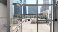 Located at 871 N Wabash Ave in Chicago's Streeterville neighborhood, this rooftop amenity floor offers a well-appointed combination of indoor and outdoor spaces set against a striking urban skyline backdrop. The expansive open-air terrace is finished with large-format porcelain tiles and furnished with contemporary modular lounge seating arranged around a central fire pit bowl, creating an inviting gathering environment. Pergola-style shade structures occupy one section of the terrace, while planters with lush greenery add organic contrast to the surrounding architecture. Glass railing panels along the perimeter preserve unobstructed panoramic views of Chicago's high-rise skyline.

The interior component features a functional kitchenette finished with speckled terrazzo-style countertops, light wood cabinetry, a matte black faucet and sink, a beverage refrigerator, and a microwave. Bold, colorful abstract artwork displayed along the upper walls adds a vibrant, curated aesthetic to the space. Floor-to-ceiling windows connect the interior visually to both the outdoor terrace and a swimming pool visible below, flooding the room with natural light. Practical access points, including a stairwell re-entry door, ensure seamless circulation throughout the building. The overall ambiance is modern, polished, and distinctly urban.