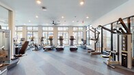 This modern fitness center is thoughtfully designed to support a full range of workout routines across multiple distinct zones. The cardio area is lined with an impressive array of treadmills, ellipticals, and stationary cycling bikes, all positioned in front of expansive floor-to-ceiling windows framed in sleek black trim. These large windows flood the space with natural light and offer views of an outdoor pool area surrounded by palm trees, creating an energizing workout environment. Large ceiling fans are installed throughout, contributing to air circulation and comfort during exercise.

The strength training zone is generously appointed with cable machines, a multi-functional Smith machine, and additional resistance equipment, all reflected in full-length mirrored walls that enhance the sense of space. A dedicated functional training studio features wood-look flooring, an "Academy"-branded performance mat, a rowing machine, wall-mounted display screen, and exposed beam ceiling details with accent lighting, offering an ideal space for high-intensity or class-based workouts.

Striped, multi-tone carpeting flows through the main training areas, adding visual depth, while smooth transitions connect each zone. Recessed lighting and large ceiling fans complete the polished, contemporary aesthetic throughout this well-appointed fitness facility.