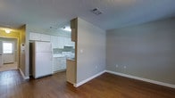 This single-floor space features an open-concept design that connects the kitchen and living area through a partial dividing wall, creating a sense of separation while maintaining an airy, connected feel. The living area is finished with warm hardwood-style flooring that extends throughout, complemented by soft gray-beige walls and crisp white baseboards that lend a clean, neutral aesthetic.

The kitchen is equipped with white cabinetry offering ample upper and lower storage, white appliances including a refrigerator and electric range, and a built-in under-cabinet range hood. A peninsula-style counter connects the kitchen to the adjacent space, providing additional workspace and a natural transition between the two areas. Recessed fluorescent lighting illuminates the kitchen zone effectively.

Both areas share a textured popcorn ceiling with visible HVAC vents, ensuring consistent climate control throughout. The living area provides a blank canvas with multiple electrical outlets positioned along the walls. The overall ambiance is straightforward and functional, offering a practical layout suited for everyday living on a single, easily navigable floor.
