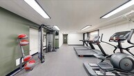 Located at 1803 Cobourg Ct in Parkville, Maryland, this single-floor amenity facility offers residents two functional spaces designed to support everyday living. The fitness center makes up the majority of the floor area and is thoughtfully outfitted with a comprehensive selection of exercise equipment. Cardio machines include multiple treadmills, an elliptical trainer, a stationary spin bike, and a rowing machine, providing a well-rounded aerobic workout experience. A multi-function cable weight machine rounds out the strength training options. The room features a neutral color palette with white and olive-toned walls, durable gray carpet flooring, and bright overhead fluorescent lighting. Windows along the wall allow natural light to filter in, offering views of the surrounding outdoor greenery. The laundry room is a compact, efficiently organized space housing multiple stacked washer and dryer units, making laundry tasks convenient for residents. The machines are coin-operated and clearly labeled, with utility infrastructure neatly integrated into the room. Together, these two spaces provide practical, well-maintained amenities that enhance the overall residential experience at this Parkville community.