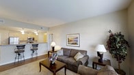 This single-floor residence offers a thoughtfully designed open-concept layout that blends comfort and practicality. The living area serves as the heart of the space, featuring warm hardwood-style flooring, neutral-toned walls, and soft ambient lighting provided by classic table lamps. A plush sofa with decorative pillows anchors the seating area, complemented by a wooden coffee table and coordinating end tables, creating an inviting atmosphere for relaxing or entertaining.

A framed landscape artwork adds a decorative focal point to the living room wall, enhancing the warm and welcoming ambiance. The space transitions naturally into the kitchen, which is accessible through an open pass-through design. The kitchen features a raised breakfast bar with bar-stool seating, pendant lighting overhead, and modern appliances including a microwave, making it both functional and stylish.

Neutral, light-colored walls and ceilings throughout lend an airy, clean feel to the interior. The open flow between the living and kitchen areas maximizes the sense of space, making the layout feel connected and efficient. Natural light filters through the space, further brightening the interior. Overall, this residence presents a balanced, cozy living environment with a practical, well-integrated floor plan.