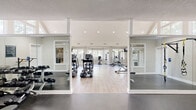 Located in Lakeland, Tennessee, this 24-Hour Fitness Gym is a thoughtfully designed fitness facility that offers a comprehensive workout experience across a single, well-organized floor. The main workout area is defined by dramatic vaulted ceilings with large, floor-to-ceiling triangular windows that flood the space with abundant natural light and provide views of the surrounding tree-lined landscape. Light wood-style flooring runs throughout the primary exercise area, contributing to a clean, modern aesthetic complemented by crisp white walls and recessed lighting. The cardio zone is generously outfitted with a row of treadmills, ellipticals, and stationary bikes, all positioned to take advantage of the expansive window views. Wall-mounted flat-screen TVs are conveniently placed for entertainment during workouts. A dedicated strength and cable machine area provides a full-body training zone, featuring multi-functional cable systems and weight benches. The free weights room is a distinct space with rubber flooring, a full-wall mirror, a tiered dumbbell rack, and a flat bench for targeted lifting sessions. An additional functional training area features wall-mounted TRX suspension systems, a flat bench, and mirrored walls, catering to bodyweight and suspension-based workouts. Throughout the facility, accessibility features and posted guidelines ensure a safe and inclusive environment for all members.