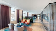 This thoughtfully designed single-floor amenity space offers a dynamic blend of bold contemporary style and functional versatility. The layout extends along a generous corridor lined with floor-to-ceiling windows dressed in sheer draping curtains, flooding the interior with abundant natural light and framing elevated views of an outdoor pool deck and surrounding cityscape.

The main lounge area is organized into distinct seating zones, each anchored by vibrant, patterned area rugs in teal, coral, and geometric motifs layered over light wood flooring. Furnishings include coral-toned rounded armchairs with yellow accent pillows, teal leather lounge chairs, striped sofas, and mixed accent seating, all arranged to encourage both social interaction and quiet retreat. Lush indoor plants are integrated throughout, adding organic warmth to the contemporary palette.

Architectural details include cove lighting along curved ceiling elements, glass-panel dividers separating zones, and a striking geometric feature wall with an integrated flat-screen display. A private meeting or focus room is finished with textured tile wallcovering, a wall-mounted desk, and distinctive circular mirror accents. Additional semi-private nooks are defined by dark panel partitions, offering more intimate gathering options within the expansive open floor plan.