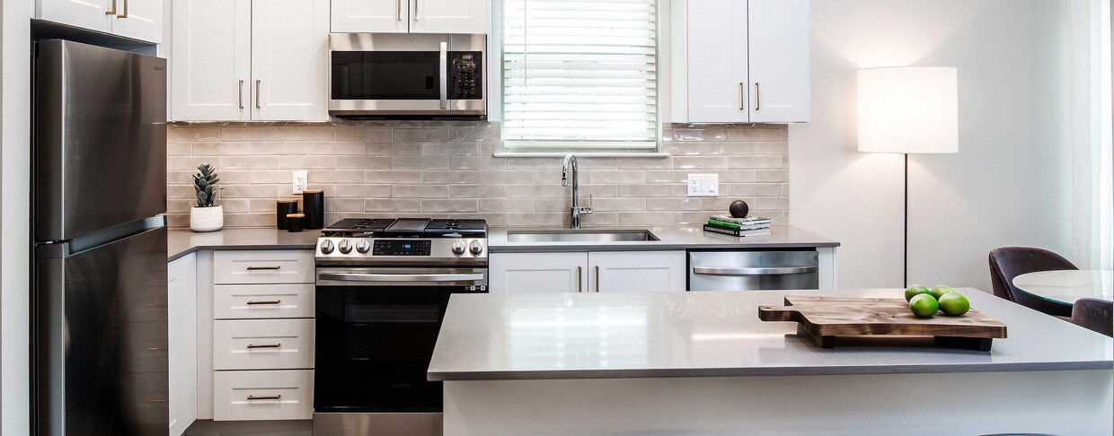 Design Collection II (South Building) Kitchen with white cabinetry, gold pulls, beige tile backsplash, grey quartz countertops, stainless steel appliances, and hard surface flooring - Avalon Somerville Station