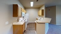 This single-floor residential unit offers a practical and well-organized layout designed for comfortable everyday living. The kitchen serves as a functional centerpiece, featuring warm wood-toned cabinetry on both sides of the galley-style corridor, light-colored countertops, white appliances including a coil-burner range, microwave, and refrigerator, and a peninsula with an integrated sink that opens toward the living and dining area. Wood-look plank flooring runs throughout the kitchen, providing a clean contrast to the neutral-toned walls. The open-concept living and dining area is carpeted and benefits from natural light through a large sliding glass door fitted with vertical blinds. A chandelier adds a subtle decorative touch to the dining space. The bedrooms are carpeted and feature neutral walls, with windows that allow natural light to filter in. At least one bedroom includes a walk-in closet with a built-in rod and shelf system. The bathroom is finished with light ceramic tile flooring, a single-sink vanity with wood cabinetry, a large mirror with overhead lighting, and a full-size soaking tub with a shower rod. The overall design is clean, neutral, and move-in ready, with a consistent warm beige color palette throughout.