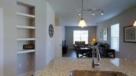 This single-floor space offers a seamlessly connected open-concept layout that integrates the kitchen, dining, and living areas into one cohesive environment. The kitchen is a standout feature, boasting granite countertops with warm, speckled tones, a modern stainless steel faucet, and pendant lighting that adds an inviting glow. Track lighting overhead ensures the space is well-lit throughout. Built-in shelving along one wall provides both functional storage and display opportunities, adding architectural character to the interior. The dining area flows naturally from the kitchen, furnished with dark wood chairs and a coordinating table, creating a defined yet open gathering space. Beyond the dining area, the living room is furnished with plush seating and anchored by a media console, offering a relaxed and comfortable atmosphere. A large window at the far end of the living room floods the space with abundant natural light, enhancing the airy feel of the open floor plan. Dark hardwood flooring runs throughout, providing a rich contrast to the light gray walls and creating a warm, modern aesthetic. Overall, the space balances contemporary design with everyday functionality.