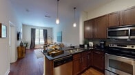 Located in Nashville's vibrant Gulch neighborhood, this single-floor residence offers a well-appointed interior defined by consistent finishes and an open, functional layout. The kitchen serves as a central hub of the space, featuring rich dark-wood cabinetry extending to notably tall ceilings, dark granite countertops, a tile backsplash, and a full suite of stainless steel appliances including a range, microwave, and dishwasher. A peninsula with bar seating provides additional prep space and a natural transition into the living area. The living room benefits from large windows dressed with floor-length drapes, allowing generous natural light to fill the space while offering views of surrounding greenery. Warm hardwood flooring runs throughout the main areas, contributing to a cohesive aesthetic. The bedroom is a calm, understated retreat with hardwood floors, large windows with slatted blinds, and ample wall space. A second bedroom offers similar comfort and natural light. The bathroom is finished with dark granite countertops, rich wood cabinetry, a large mirror, and a combined tub and shower, maintaining the design continuity found throughout the residence. Overall, the space presents a modern urban aesthetic with warm, quality finishes.