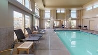 Located at 3304 E 44th Ave in Spokane, Washington, this single-floor indoor aquatic facility offers a well-appointed and functional environment centered around a generously sized lap pool. The pool area is defined by soaring ceiling heights and an expansive bank of windows fitted with wood-toned horizontal blinds, allowing abundant natural light to flood the space while providing views of an outdoor patio area with umbrellas and greenery beyond.

The pool surround features a wide, tiled deck with ample room for circulation, lined with modern lounge chairs and side tables that create comfortable areas for relaxation before or after swimming. The neutral palette of warm beige and sage tones throughout the walls and tile wainscoting lends the space a calm, resort-like atmosphere.

Adjacent to the main pool deck, a transitional seating area near the entrance provides additional lounge chairs and a wooden bench, offering a welcoming space for visitors. Dual stainless steel drinking fountains mounted at different heights are conveniently located along the corridor, reflecting the facility's attention to accessibility and practical amenities. The overall design balances functionality with comfort, making this a well-equipped aquatic space suited for both recreational and fitness use.