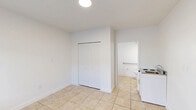 This 230 square foot studio apartment, located at 935 W Okeechobee Rd in Hialeah, FL, offers a compact yet functional living space. The single-floor layout comprises a main living area and a separate bathroom. Throughout the apartment, beige tile flooring provides a neutral base, complemented by crisp white walls that enhance the sense of space.

The main living area is equipped with a window air conditioning unit, ensuring climate control in the Florida heat. A large window allows natural light to flood the space, while horizontal blinds offer privacy when needed. The kitchenette, though minimal, includes a small countertop with a sink and a compact refrigerator, suitable for basic food preparation.

The bathroom, while modest in size, features essential amenities including a toilet and a safety grab bar. A small window in the bathroom provides ventilation and natural light. The apartment's entrance is secured with a modern electronic lock system, adding a touch of contemporary convenience to the space.

Storage options appear limited, with a single closet visible in the images. The overall design prioritizes functionality in a small footprint, making efficient use of the available square footage. This studio would be suitable for a single occupant seeking a no-frills living space in an urban setting.