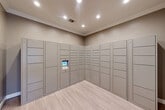 Located at 555 Spring Park Center Blvd in Spring, Texas, this dedicated package locker room is a well-appointed amenity space designed for secure and convenient parcel management. The room is finished with light wood-look flooring that runs throughout, complemented by warm taupe-toned walls and crisp white crown molding that lend a polished, cohesive aesthetic.

The focal point of the space is a floor-to-ceiling smart parcel locker system that wraps along two walls in an L-shaped configuration. The lockers are finished in a neutral gray tone and vary in compartment size to accommodate packages of different dimensions. A centrally mounted digital touchscreen interface provides residents with a streamlined, self-service retrieval experience.

Recessed lighting installed in the ceiling provides bright, even illumination throughout the room, ensuring clear visibility at all times. A large-scale abstract artwork with warm earth tones, creams, and muted blues is mounted on the wall near the entry, adding an artistic and welcoming touch to an otherwise functional space.

The entry corridor leading to the room features the same light flooring and taupe walls, with a doorway opening to an adjacent area with natural light visible through window blinds. Overall, the space balances practical utility with thoughtful design details, making it a refined and efficient amenity.