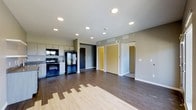 This single-floor space showcases a thoughtfully designed open-concept layout that seamlessly connects the kitchen and living areas. The kitchen is equipped with black appliances, including a refrigerator, range, and built-in microwave, complemented by light-toned cabinetry that provides ample storage. The countertops and cabinetry offer a clean, functional workspace suited for everyday use.

Durable hardwood-style flooring runs throughout the space, transitioning between two complementary tones that add visual interest and warmth to the interior. The recessed lighting installed across the ceiling ensures even, modern illumination throughout the open area.

The neutral gray walls and crisp white trim create a contemporary aesthetic that feels both clean and inviting. Ceiling vents indicate a centralized HVAC system for year-round climate control. Multiple doorways visible from the main living area lead to additional rooms, suggesting a well-connected floor plan with clear separation between living and private spaces. A dark accent wall near the entry door adds a bold design touch, grounding the space with contrast. Overall, the unit presents a modern, move-in-ready interior with practical finishes and a cohesive design sensibility.